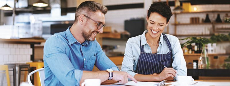 Employer points to a clipboard while he and employee have coffee.