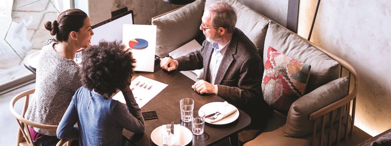 Two women and a man review charts and graphs following a meal in a restaurant.